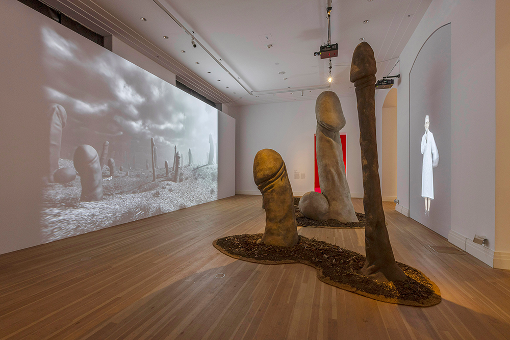 Skulpturen in Form von übergroßen Phallusfiguren in einem Ausstellungsraum mit zwei projizierten Schwarz-weiß-Filmen an den Wänden