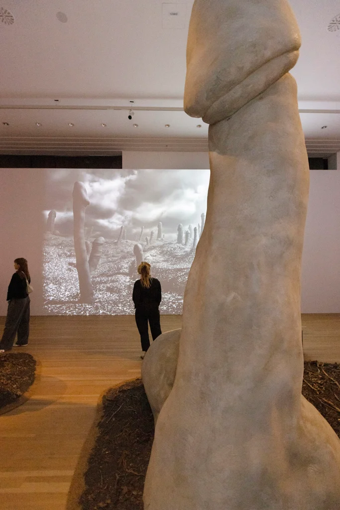 Große Skulptur in Form eines Phallus in einem Ausstellungsraum mit zwei Personen und einer schwarz-weißen Projektion von ähnlichen Skulpturen an der Wand