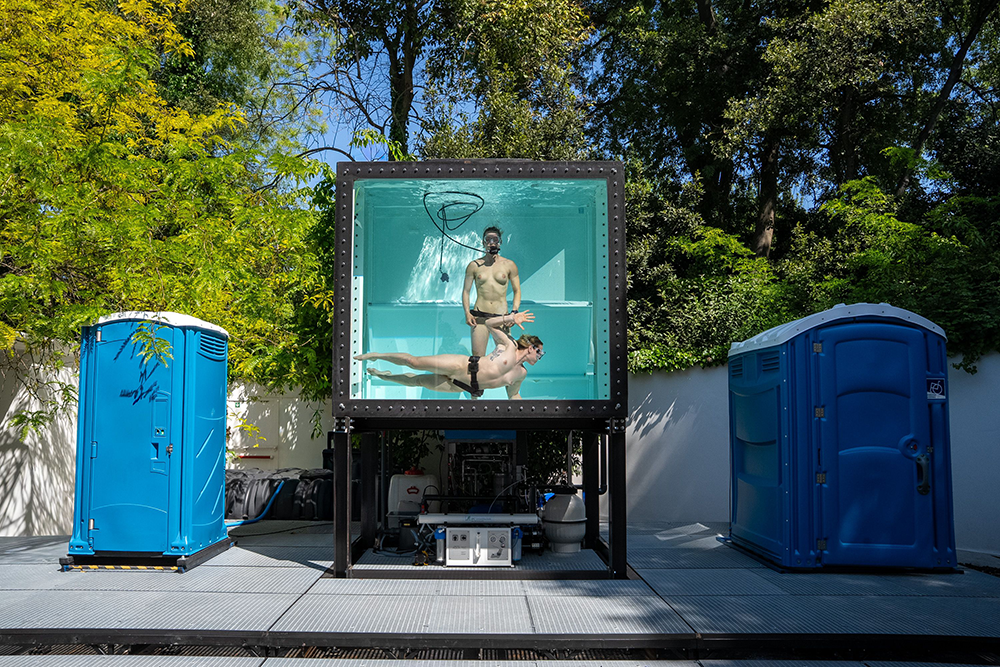 Zwei Menschen in Badebekleidung schwimmen in einem großen, transparenten Wasserbecken, das auf einem schwarzen Gestell steht, umgeben von zwei blauen mobilen Toiletten und Bäumen im Hintergrund