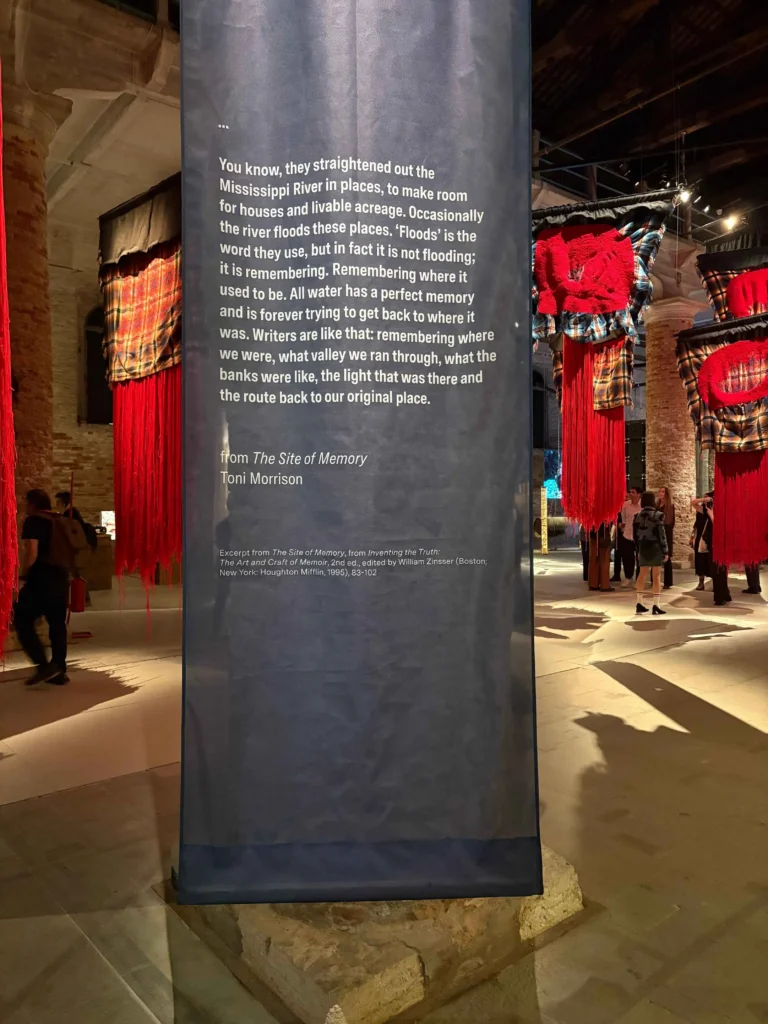 Dunkelblaues Banner mit weißem Textzitat von Toni Morrison in einer Ausstellung mit rot-schwarzen Kleidungsstücken im Hintergrund.