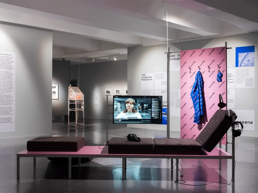 Ausstellungsszene mit einem pinkfarbenen Tisch, darauf ein braunes Polster mit verstellbarer Lehne, im Hintergrund ein Monitor mit einem Gesicht und Textwänden.
